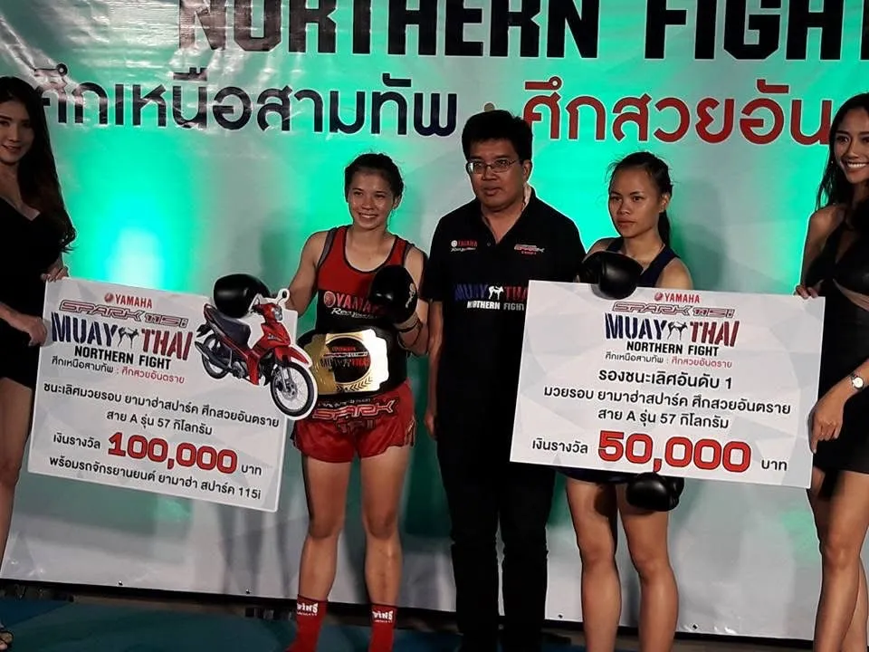 Kru Cream in red holding the championship belt beside the 100,000 baht prize board